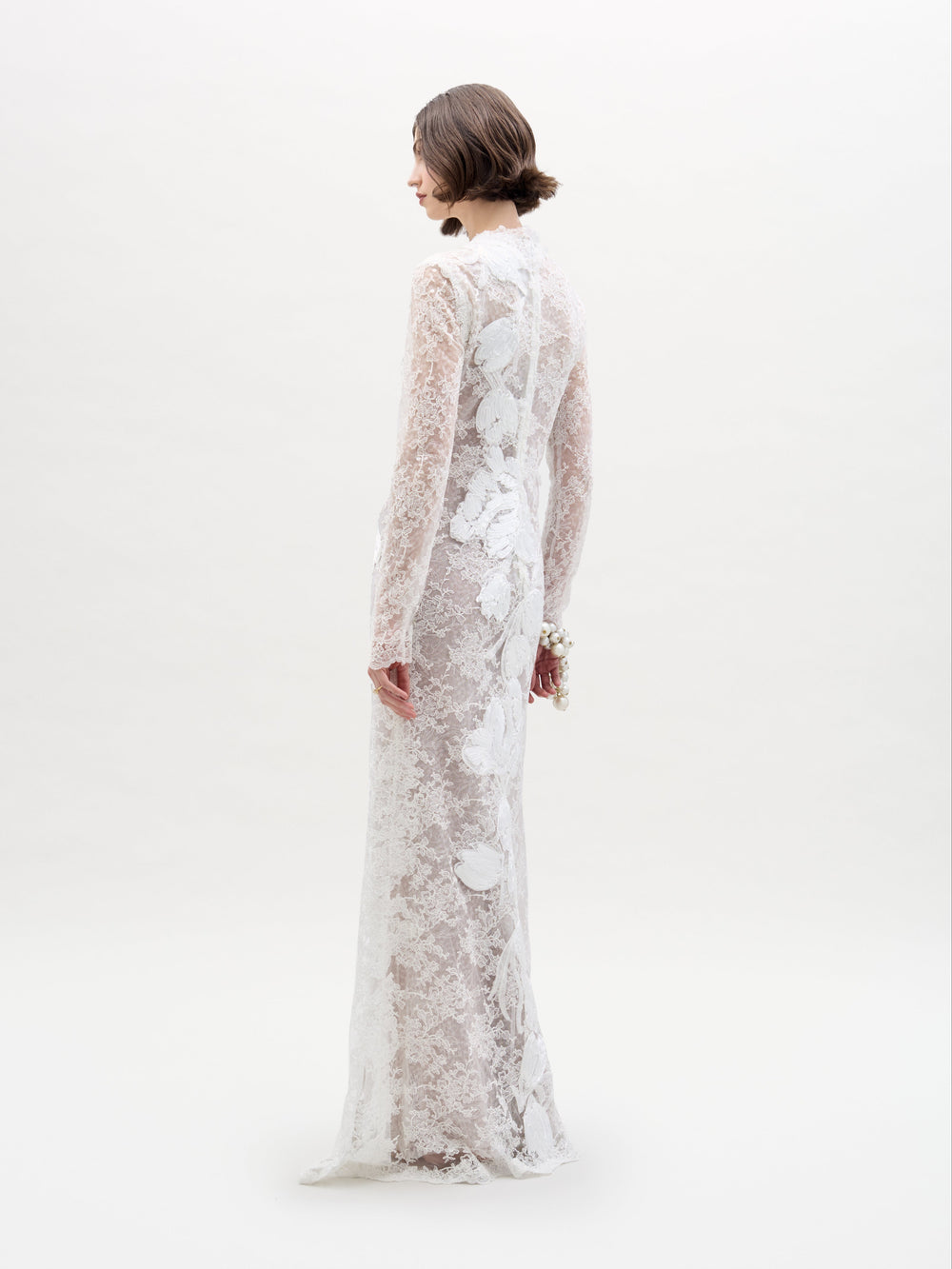A woman stands against a plain background wearing the SVT DESIGNS Dava Dress, showcasing its long-sleeved, floor-length design. The dress is white with a beautiful floral lace pattern as she faces away, highlighting the back and side.