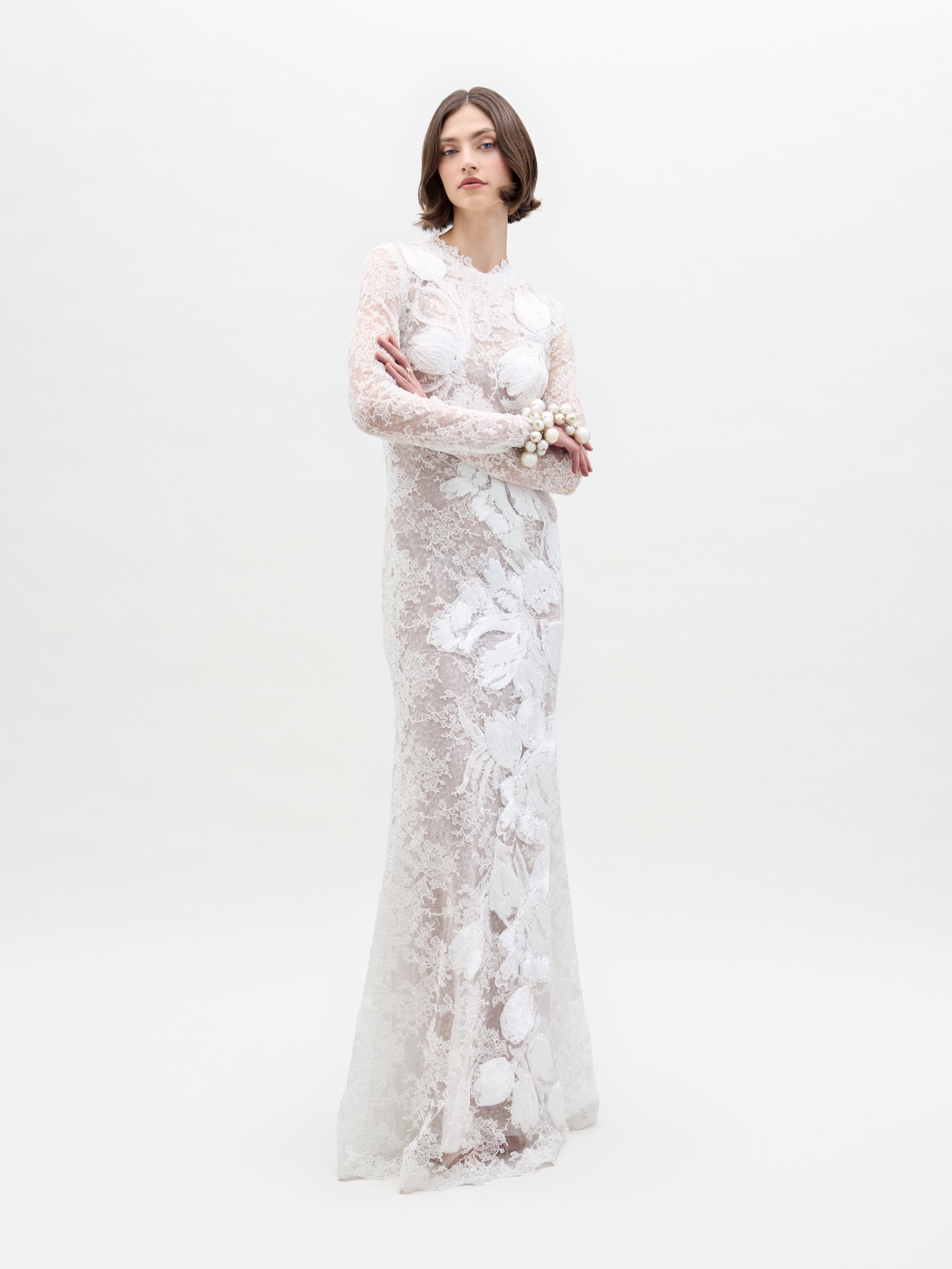 A person is wearing the Dava Dress, a floor-length, long-sleeved lace gown featuring floral patterns from SVT DESIGNS. They stand against a plain white background with short hair, holding their right arm with their left hand.