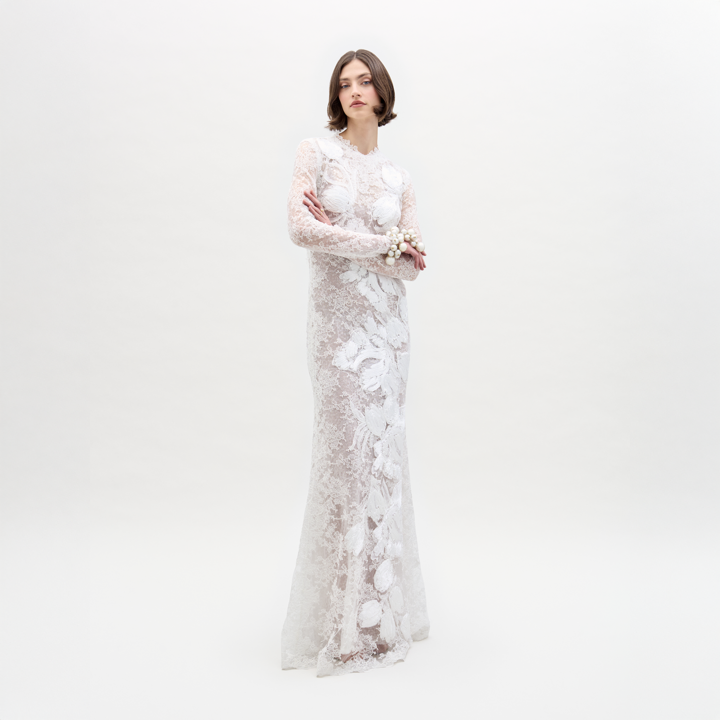 A woman with short brown hair and a neutral expression stands against a white background, arms crossed, wearing the long-sleeve lace Dava Dress by SVT DESIGNS in a column silhouette, accessorized with a floral bracelet.