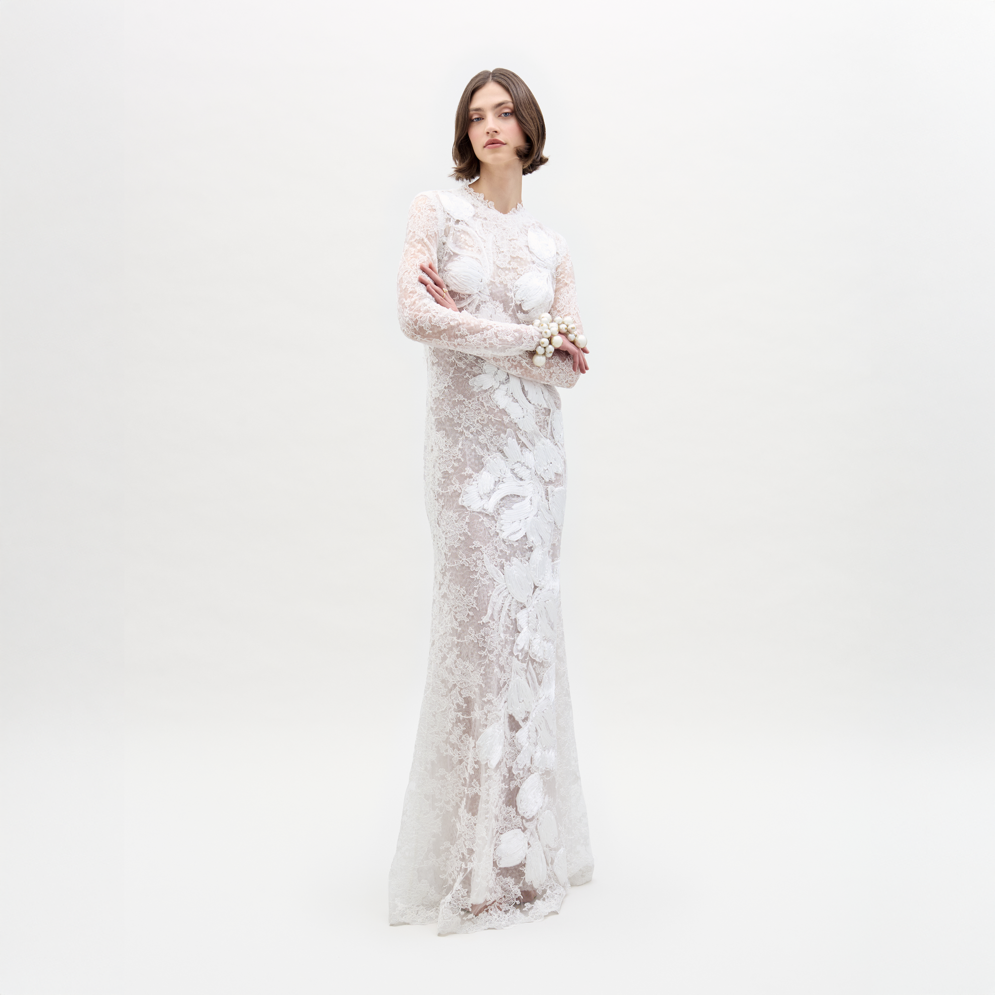 A woman with short brown hair and a neutral expression stands against a white background, arms crossed, wearing the long-sleeve lace Dava Dress by SVT DESIGNS in a column silhouette, accessorized with a floral bracelet.