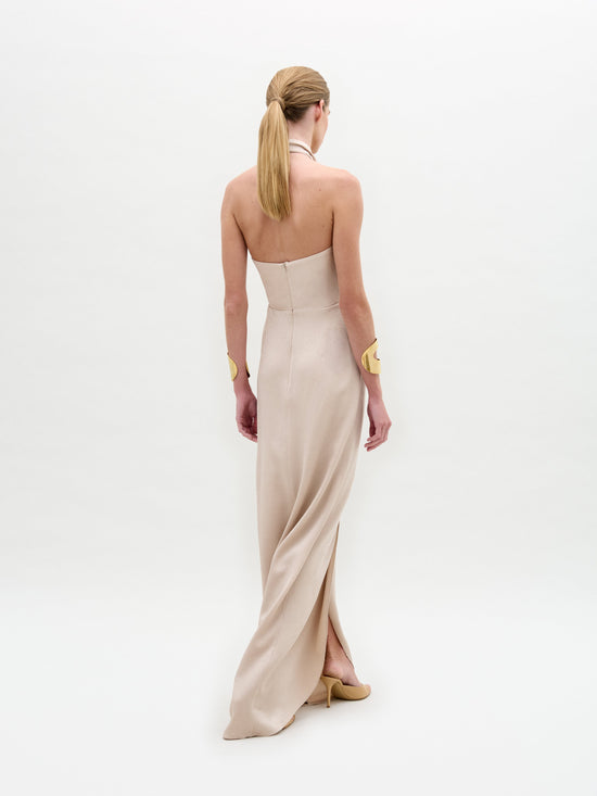 A person with long light brown hair, styled in a low ponytail, stands with their back to the camera, adorning a strapless, floor-length beige Davinia Dress by SVT DESIGNS and wearing gold-colored bracelets against a plain white background.