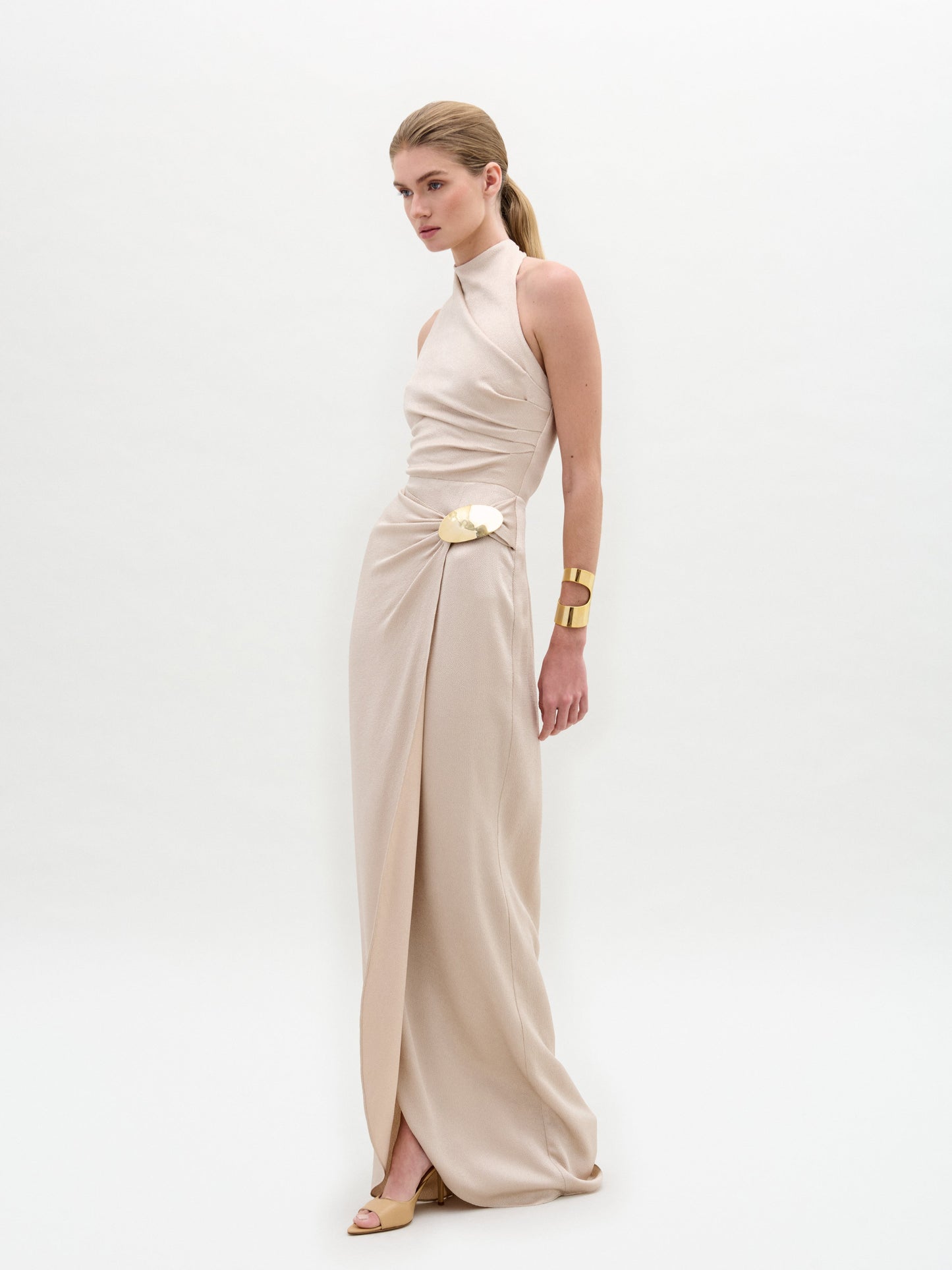 A woman wears the Davinia Dress by SVT DESIGNS, a long beige sleeveless gown with a high neckline and a floral waist accessory. She pairs it with chunky gold bracelets and beige open-toe heels against a plain white background.