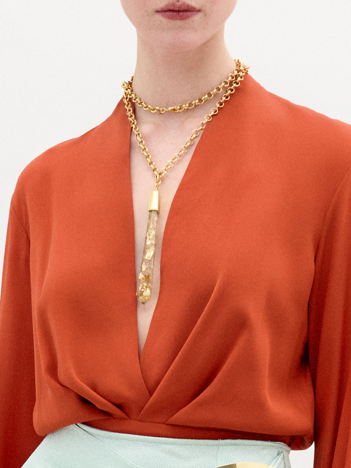A woman wearing the long-sleeve rust-colored Delfina Blouse, a light blue skirt with a bold metallic waist accent, and layered gold necklaces stands against a plain white background.