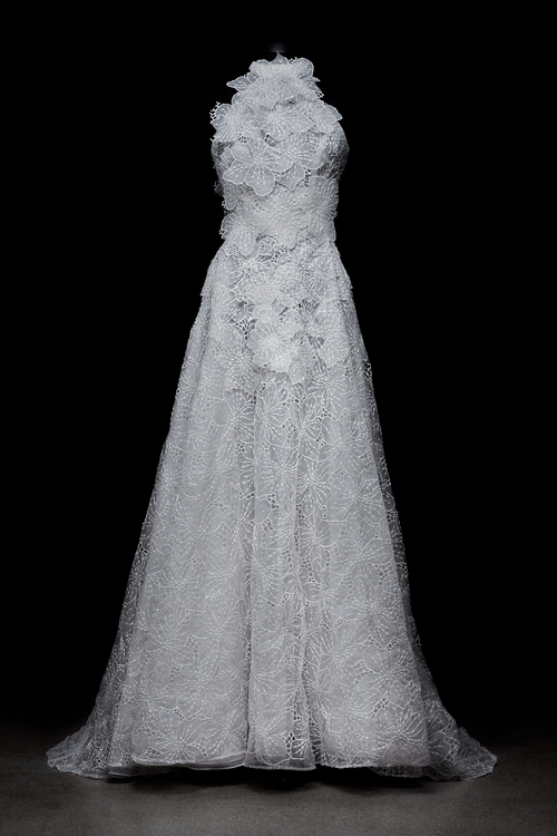 A woman in the Dorothy Dress Pearl, an exclusive white lace wedding gown with a high neck and sleeveless design, stands against a light background.
