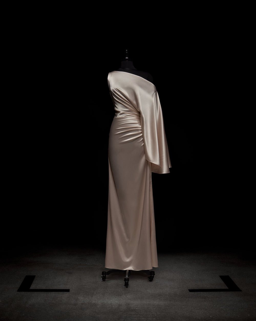The Dotty Dress by SVT DESIGNS is showcased on a mannequin against a dark backdrop. This elegant champagne-colored satin gown features an asymmetrical, off-the-shoulder design with a floor-length silhouette and draped fabric on one side.