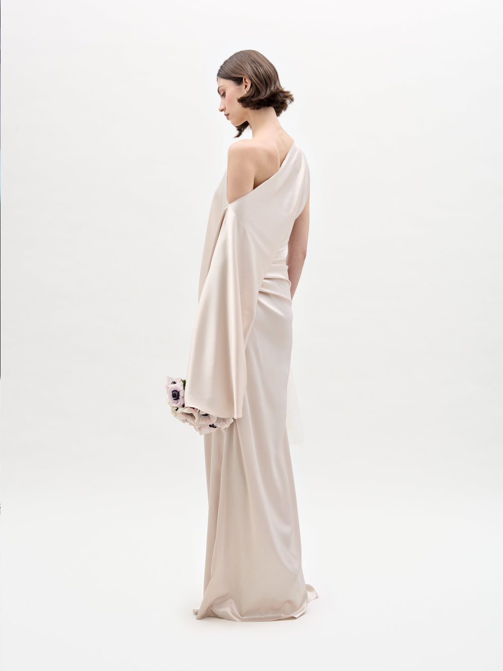 A woman in an elegant asymmetrical beige Dotty Dress from SVT DESIGNS stands against a white background. She holds a floral-detailed clutch and has a short bob hairstyle, looking slightly down to her left.