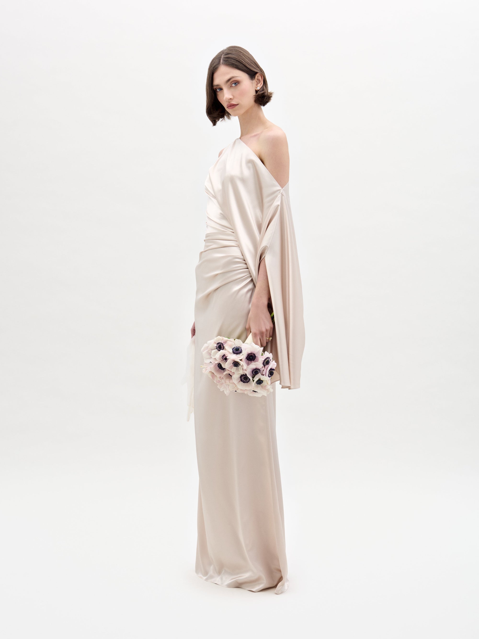 A woman wearing the SVT DESIGNS Dotty Dress, an elegant ivory satin off-the-shoulder gown, stands against a plain white background. She holds a bouquet of white and dark-centered flowers, with her dark hair styled in a short bob.