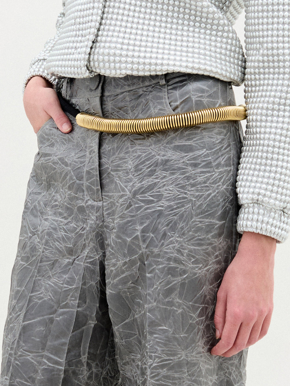 A person stands in a white textured zip-up jacket and gray Emine Pant with a gold belt against a plain white background.