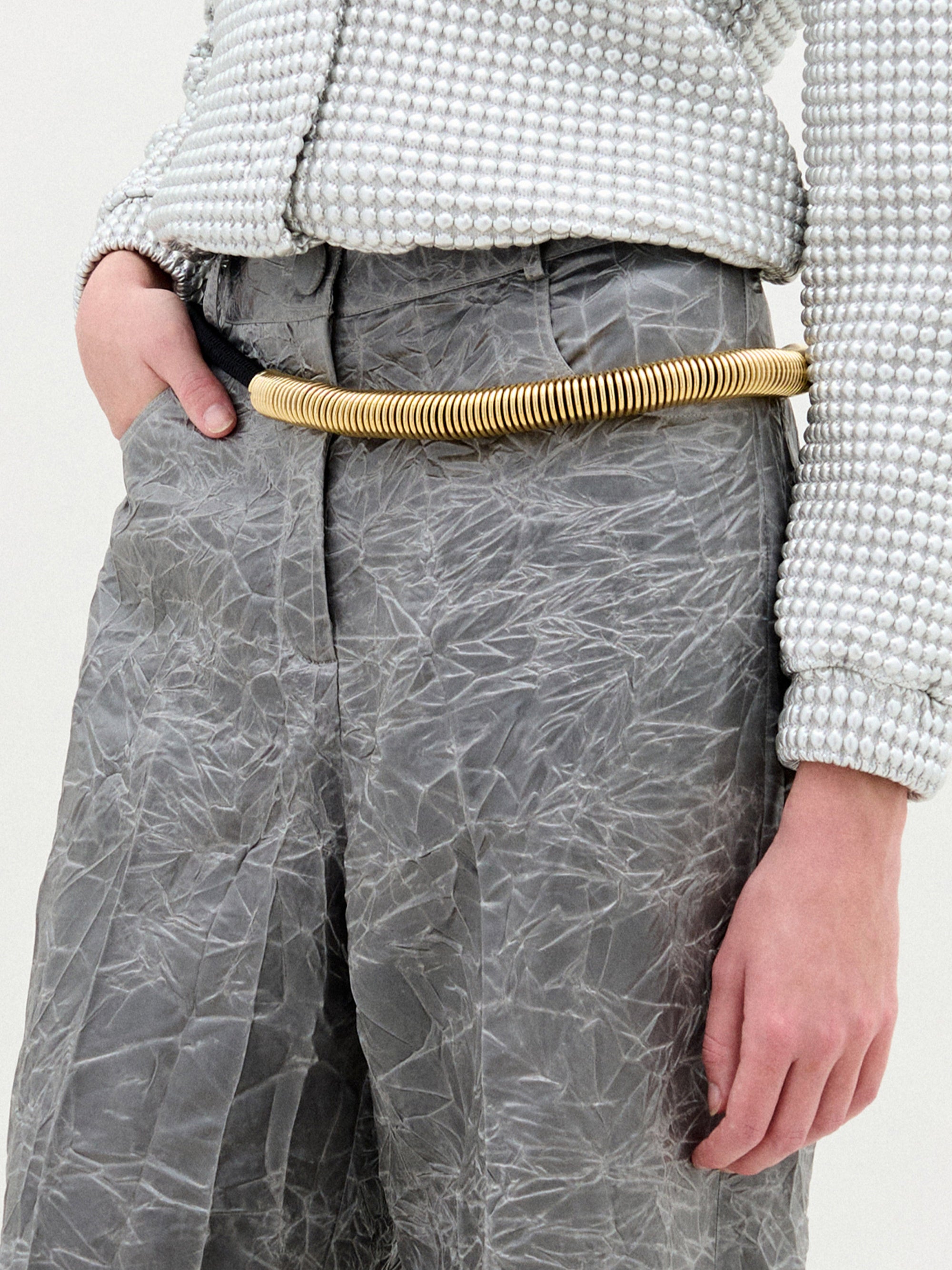 A person stands in a white textured zip-up jacket and gray Emine Pant with a gold belt against a plain white background.