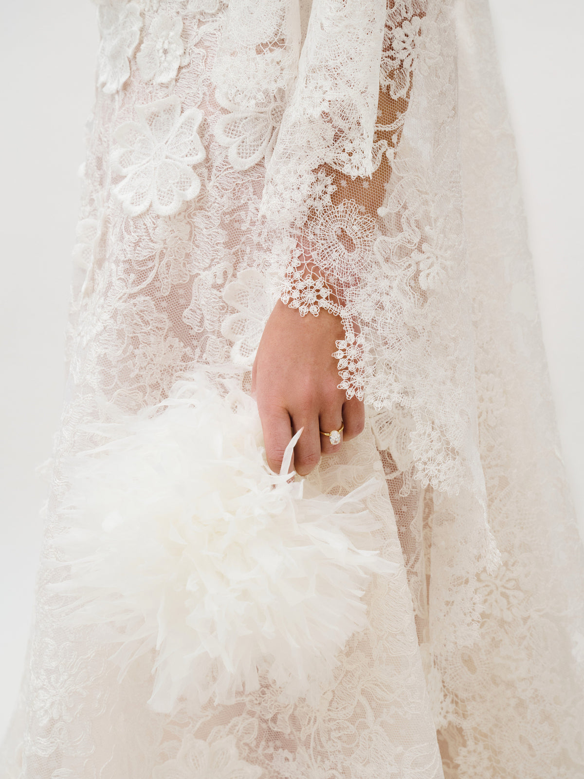A person wearing the Danita Dress, made from exquisite Italian fabric by Colombia Atelier and adorned with intricate white floral lace, holds a fluffy white feathered accessory. Their hand displays a ring, while the light, neutral background complements the detailed elegance of the dress.