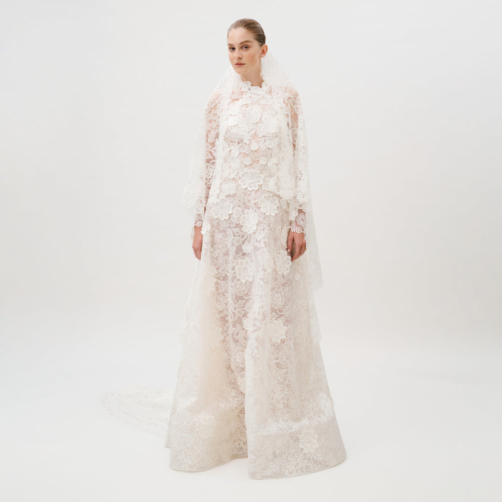 Dressed in the Danita Dress—a long, white, lace wedding gown adorned with floral patterns and crafted from exquisite Italian fabric by Colombia Atelier—a woman stands against a plain white background. Her timeless bridal look is completed with a matching lace veil.