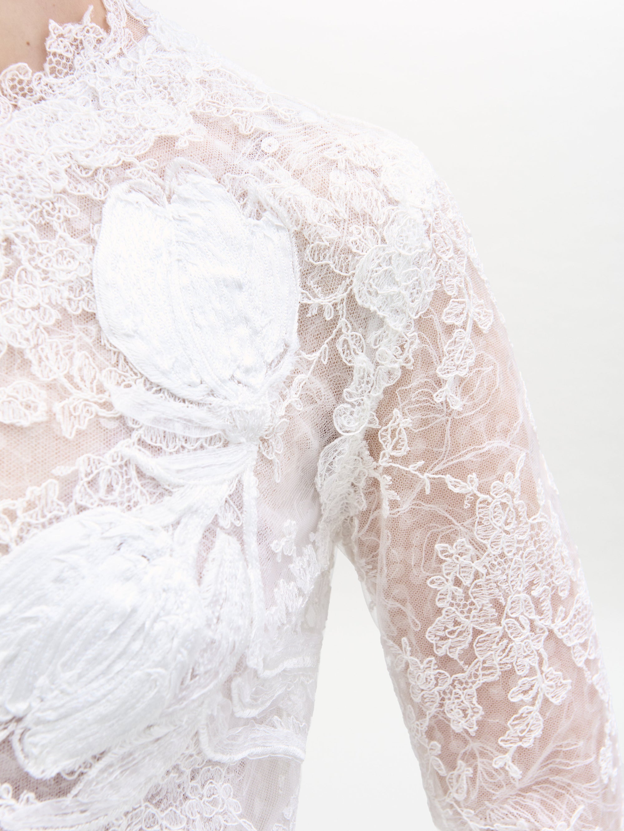 Close-up of the Dava Dress by SVT DESIGNS, crafted from sheer white lace with floral patterns, intricate embroidery, and larger white floral appliqués. The delicate lace covers the shoulder and chest area.