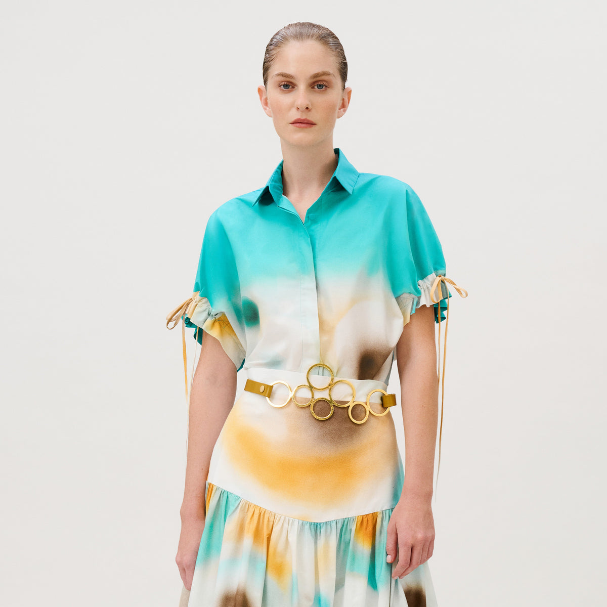 The Dixie Shirt and matching skirt in 100% cotton feature a teal, white, and yellow abstract print, drawstring sleeves, and a waist belt decorated with interlocking gold-tone metal rings.
