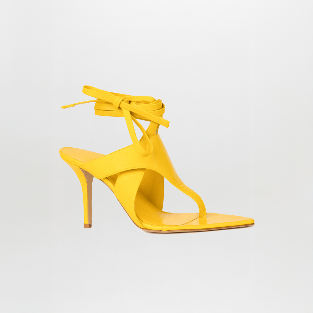 The Domenico Heels Limoncello by SilviaTcherassi are yellow Italian leather heeled sandals with a thong and square toe, ankle wrap laces, and a stiletto heel—the minimal design pops against a plain white background.