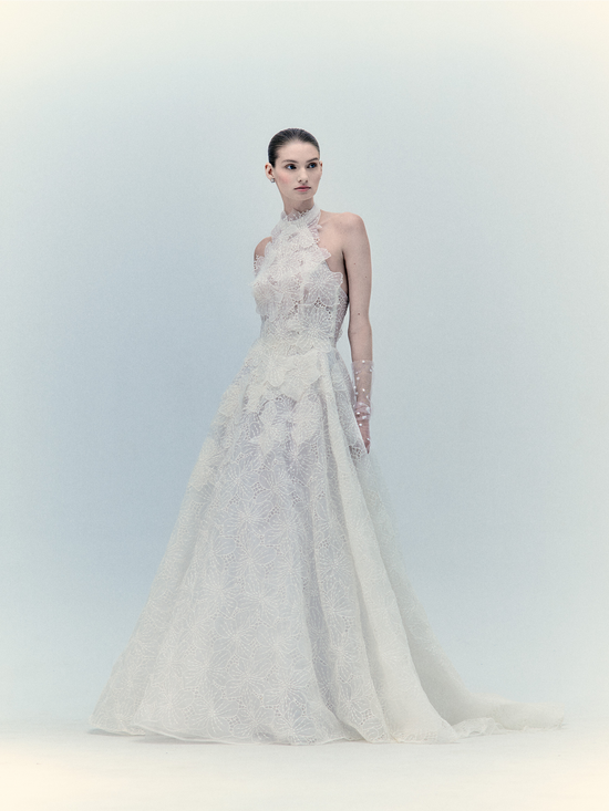 A woman in the Dorothy Dress Pearl, an exclusive white lace wedding gown with a high neck and sleeveless design, stands against a light background.