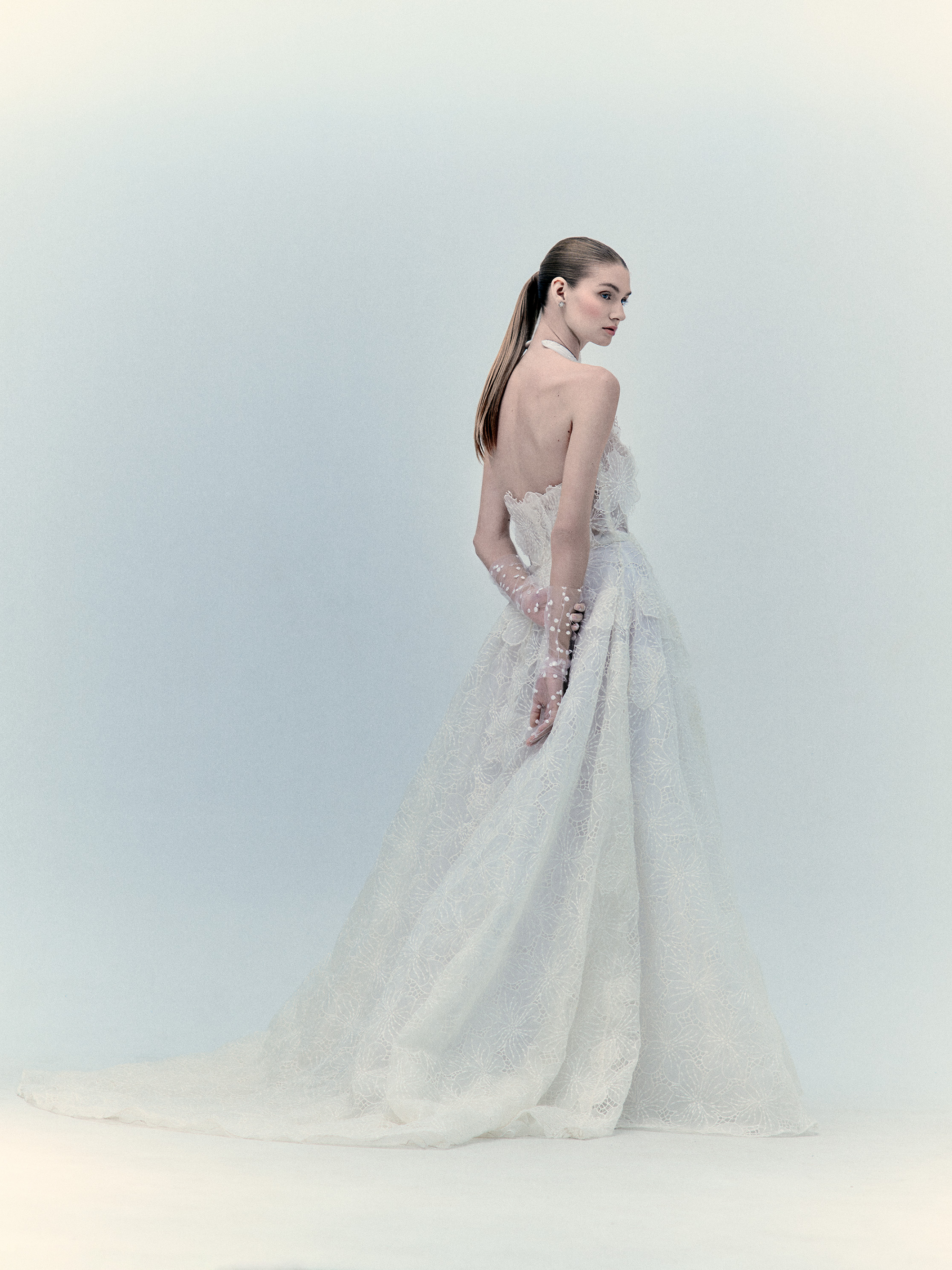 A woman stands in profile facing left, wearing the SVT DESIGNS Dorothy Dress—a strapless white maxi gown with a long, textured skirt and sheer gloves. Her straight hair is pulled back, set against a plain, light-colored background.