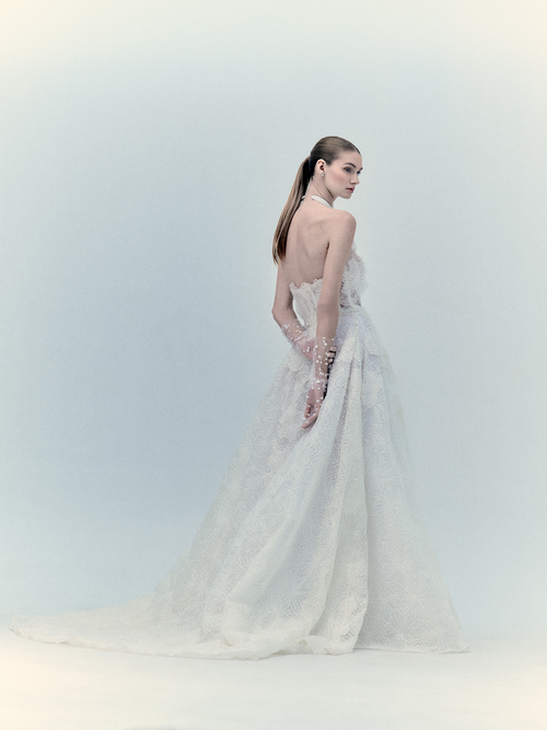 A woman in the Dorothy Dress Pearl, an exclusive white lace wedding gown with a high neck and sleeveless design, stands against a light background.