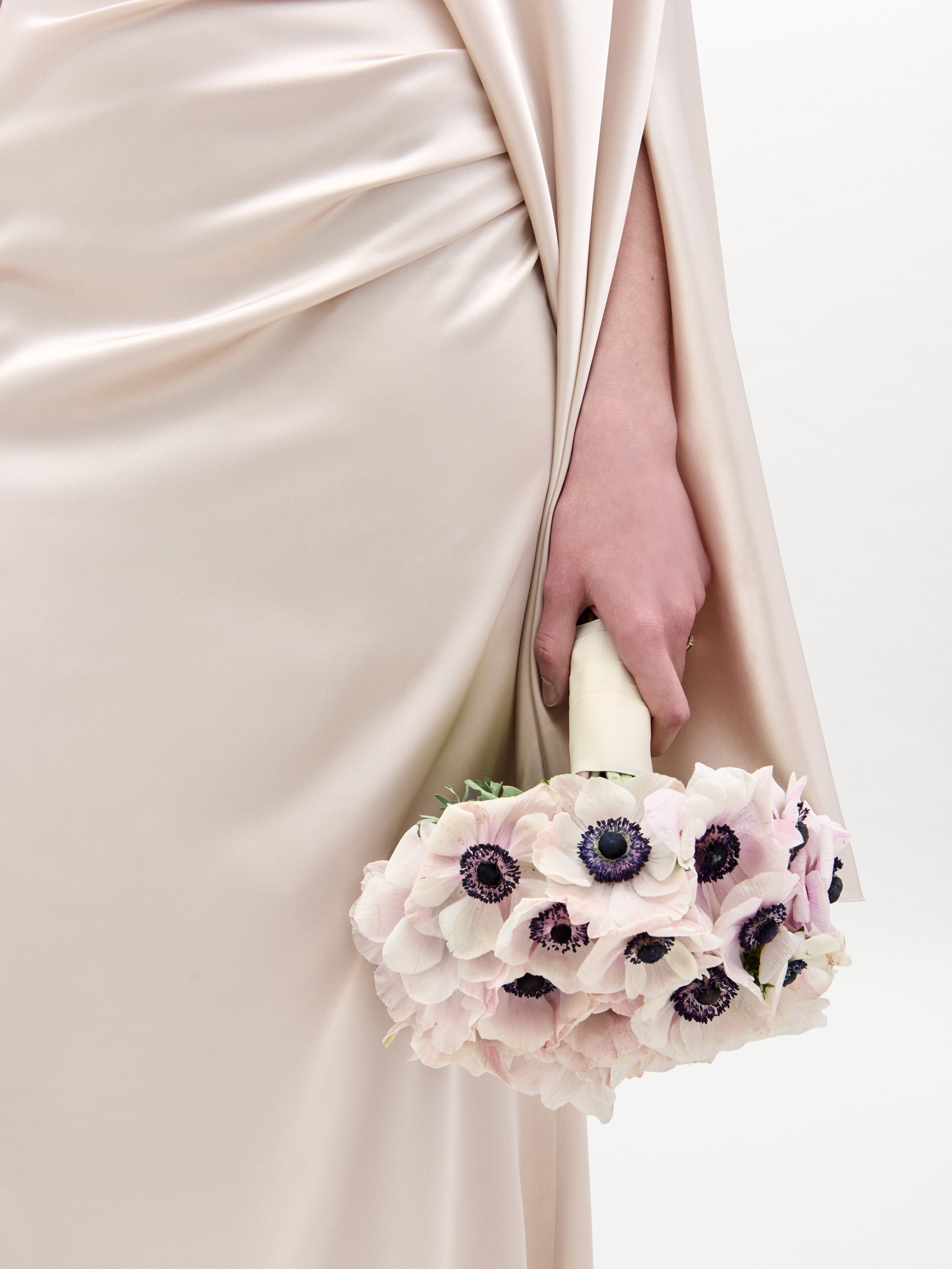 A person wearing SVT DESIGNS' Dotty Dress, a silky cream-colored gown with draped fabric and soft folds, holds a bouquet of light pink anemones with dark centers against a plain white background.