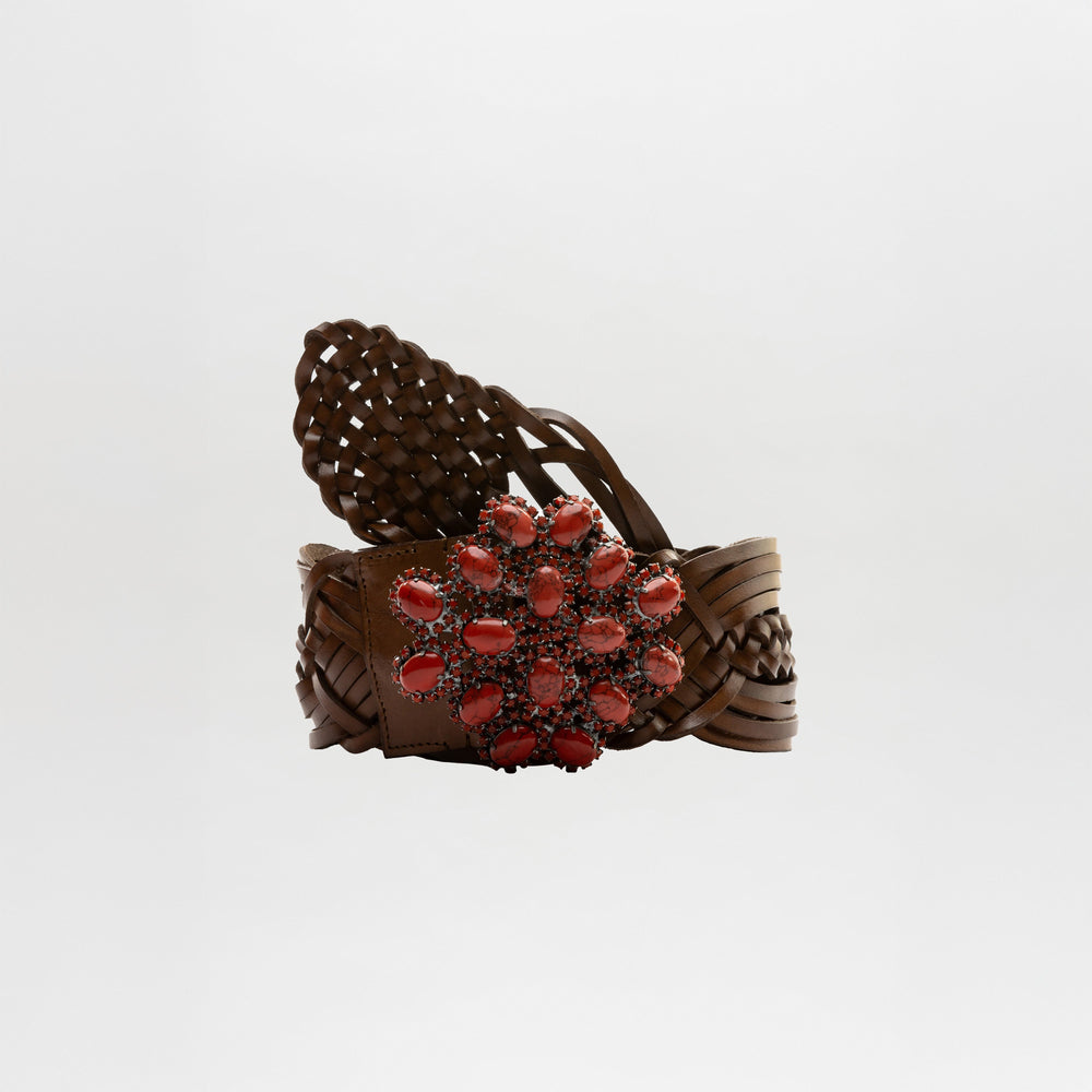 The Eines Belt by SVT DESIGNS is a wide braided Italian leather belt with a large floral buckle decorated with red oval stones. It features a plain, light-colored background and is made in Italy.