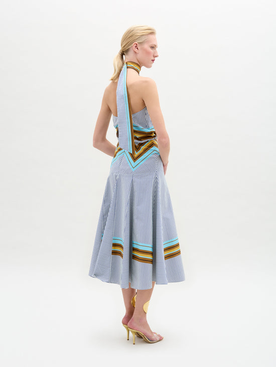 A blond person poses in profile against a plain background, wearing the Elara Dress by SVT DESIGNS. This halter neck midi dress is crafted from fine Italian fabric with vertical stripes and multicolored bands, features an A-line skirt with a long scarf-like back detail, and is paired with high heels.