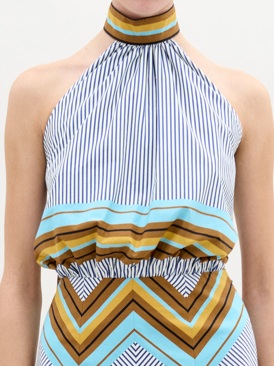 A person is wearing the Elara Dress by SVT DESIGNS. This halter, high-neck midi dress features blue and white vertical stripes on the top and an A-line skirt with a geometric design in brown, turquoise, and beige diagonal shapes, made from exquisite Italian fabric.