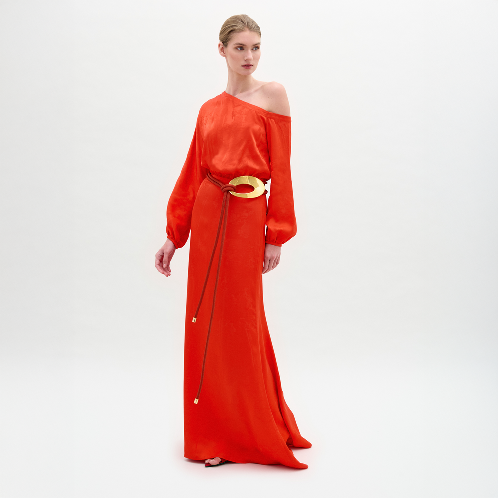A woman poses in the SVT DESIGNS Elga Dress, featuring a long, bright red one-shoulder maxi silhouette with long sleeves and a gold 3D cord belt, set against a plain white background as she looks to the side.