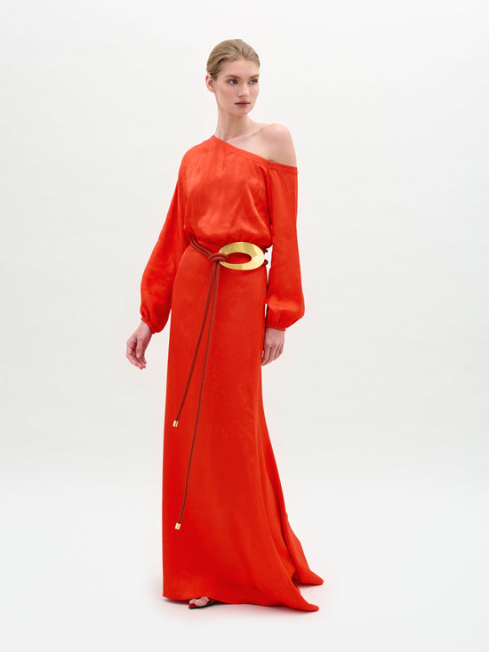 Against a white background, a person models the SVT DESIGNS Elga Dress, a striking red maxi with an asymmetric neckline and long sleeves. Made from luxurious Italian fabric, it's styled with a wide gold belt featuring two hanging ropes as they gaze off to the side.