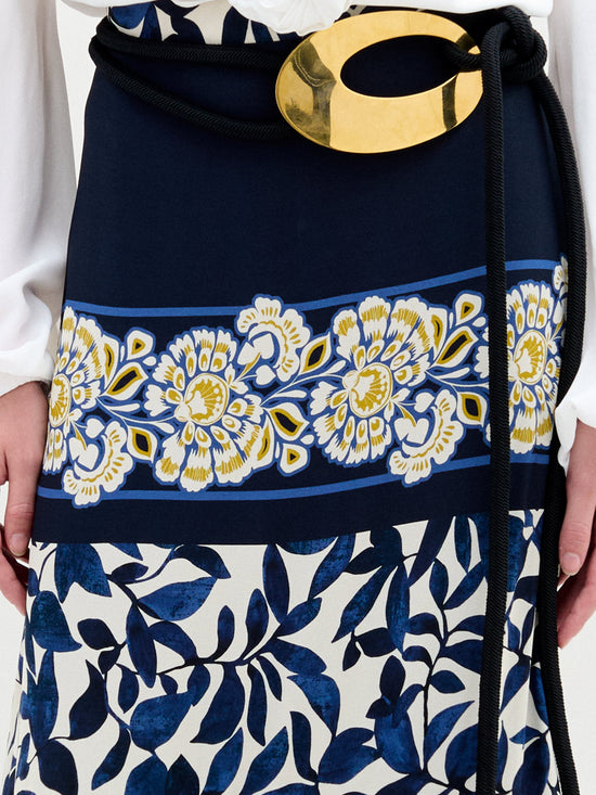 A close-up displays the Elliot Skirt by SVT DESIGNS, an asymmetrical maxi skirt in a blue and white floral pattern with yellow accents, crafted from fine Italian fabric. It's tied with a black rope belt featuring a large gold oval buckle. The person pairs it effortlessly with a stylish white top.