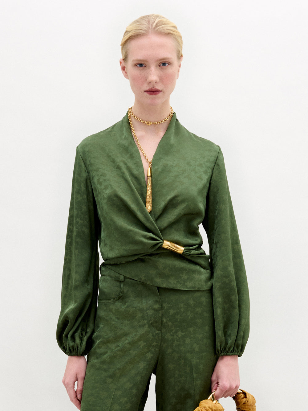 A person with light skin and blonde hair pulled back wears the SVT DESIGNS Elvie Blouse, a green long-sleeved wrap top with matching pants, standing against a white background, accessorized with gold jewelry and a gold waist detail.
