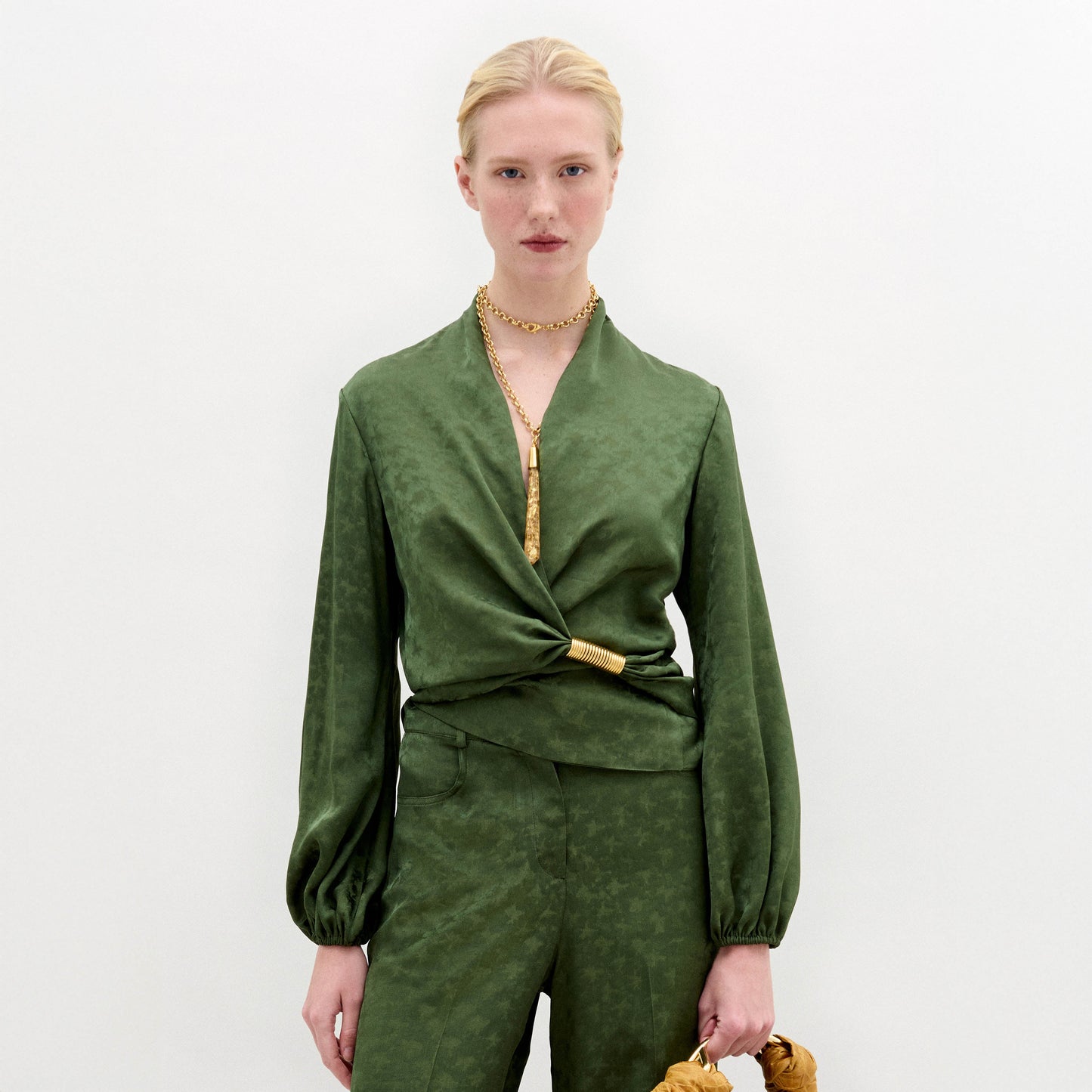Person standing against a plain background wearing the green Elvie Blouse, a long-sleeved wrap top with wrap-around ties, matching pants, accessorized with a gold necklace and holding a gold handbag.