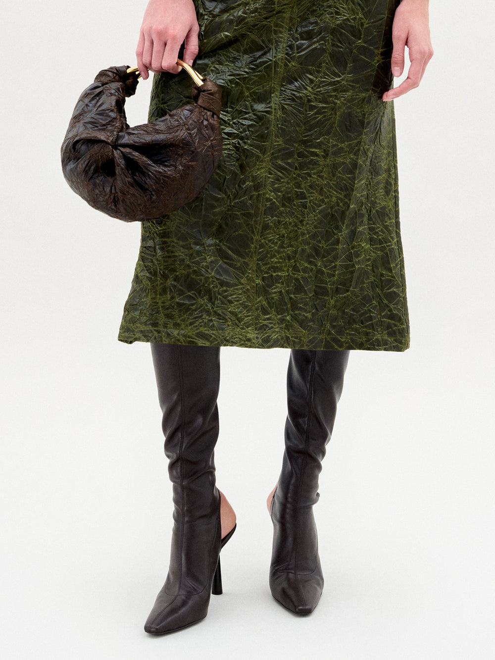 A person in a textured green skirt and SVT DESIGNS Ember Boots holds a dark brown, crescent-shaped handbag with a short handle. The plain, light-colored background highlights the Italian leather boots.