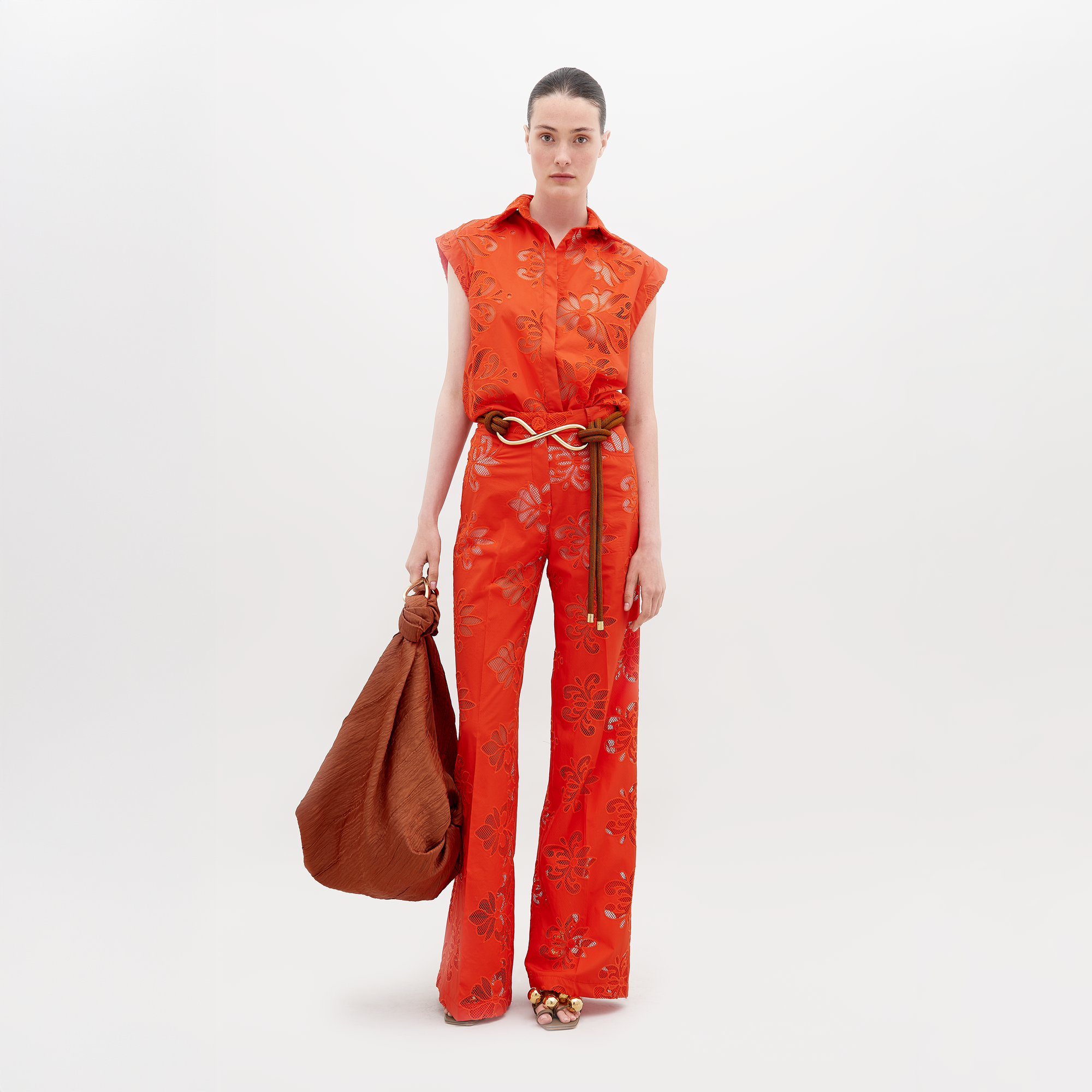 A person stands against a plain background in the Emine Pant by SVT DESIGNS, featuring a sleeveless red floral pattern, wide-leg silhouette, and braided belt. They hold a large brown bag, wear open-toed shoes, have their hair pulled back, and display a neutral expression.