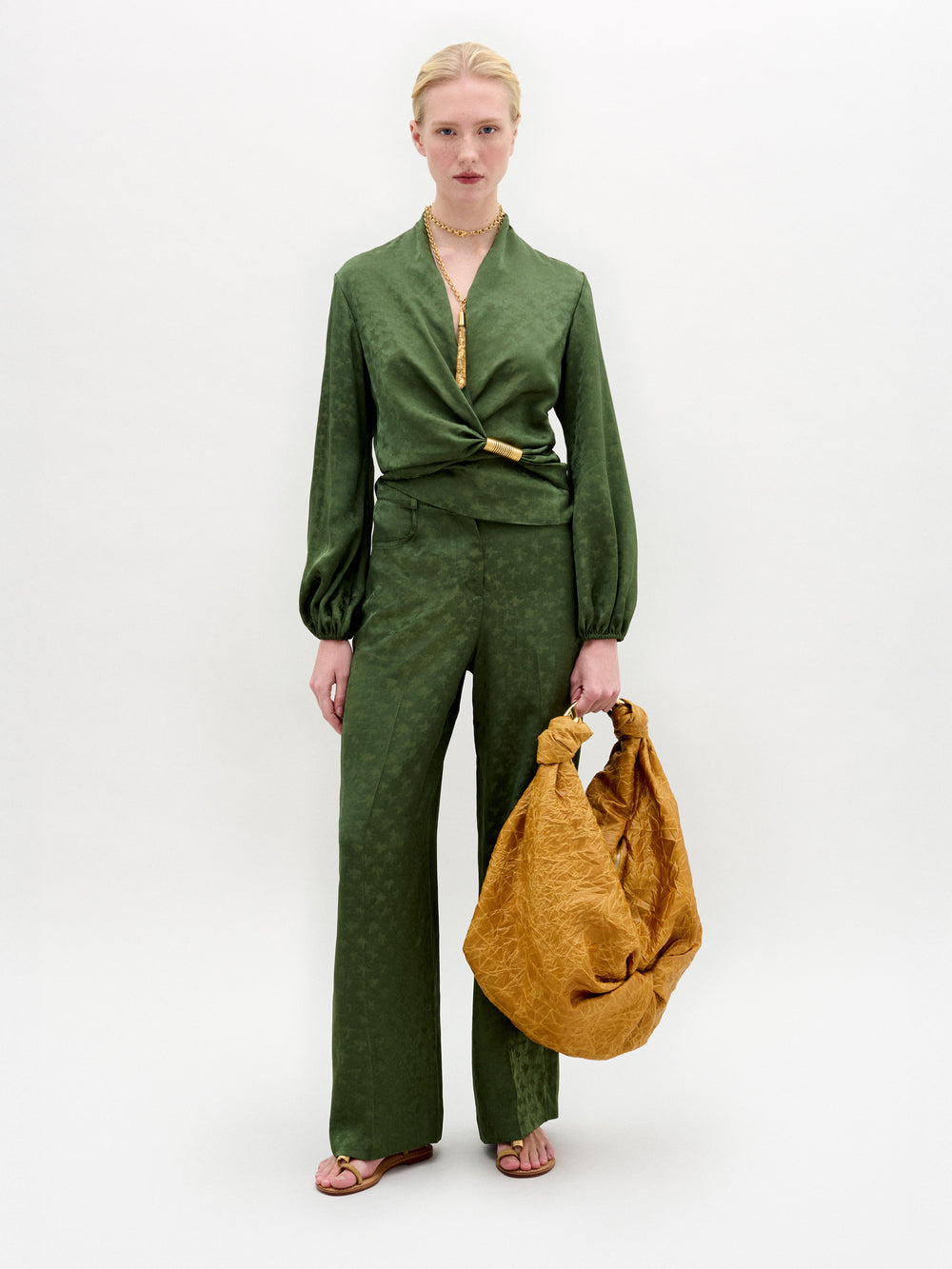 A person stands against a plain background wearing the Emine Pant by SVT DESIGNS in green, paired with a matching wrap tie blouse, brown sandals, and holding a large crumpled brown bag.