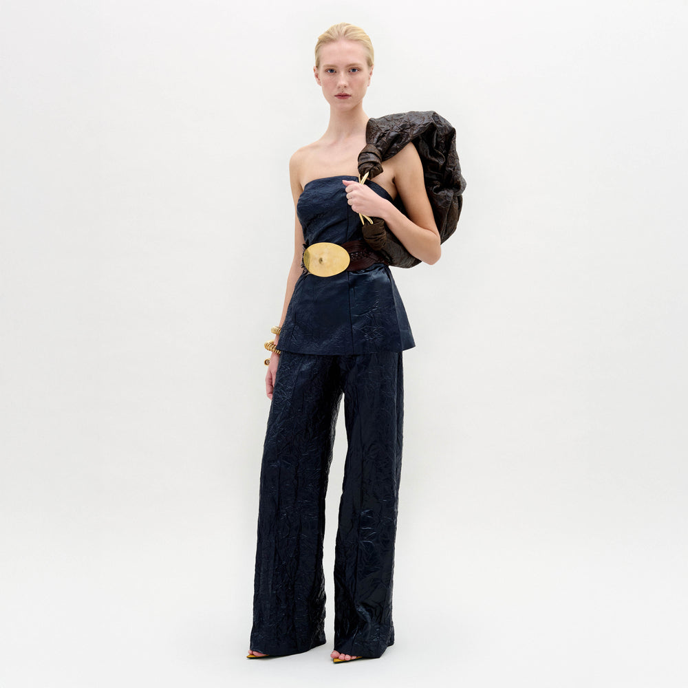 A woman poses against a white backdrop wearing the SVT DESIGNS Emine Pant, a high-waisted, wide-leg silhouette in navy blue, paired with a matching strapless top, gold belt and bangles, and a dark textured shoulder bag.