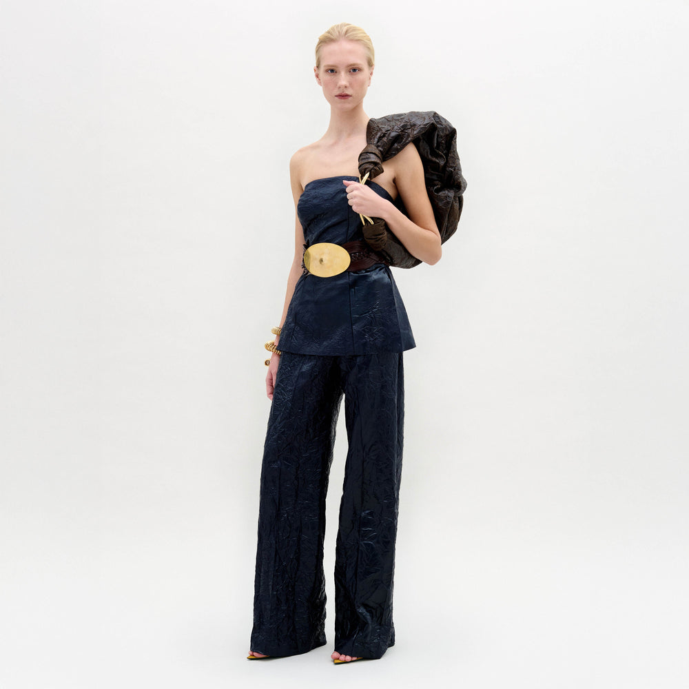 A woman stands against a white background wearing the Emine Pant—a high-waisted, wide-leg silhouette—paired with a dark strapless top, gold belt, bangles, and a textured bag slung over her shoulder.