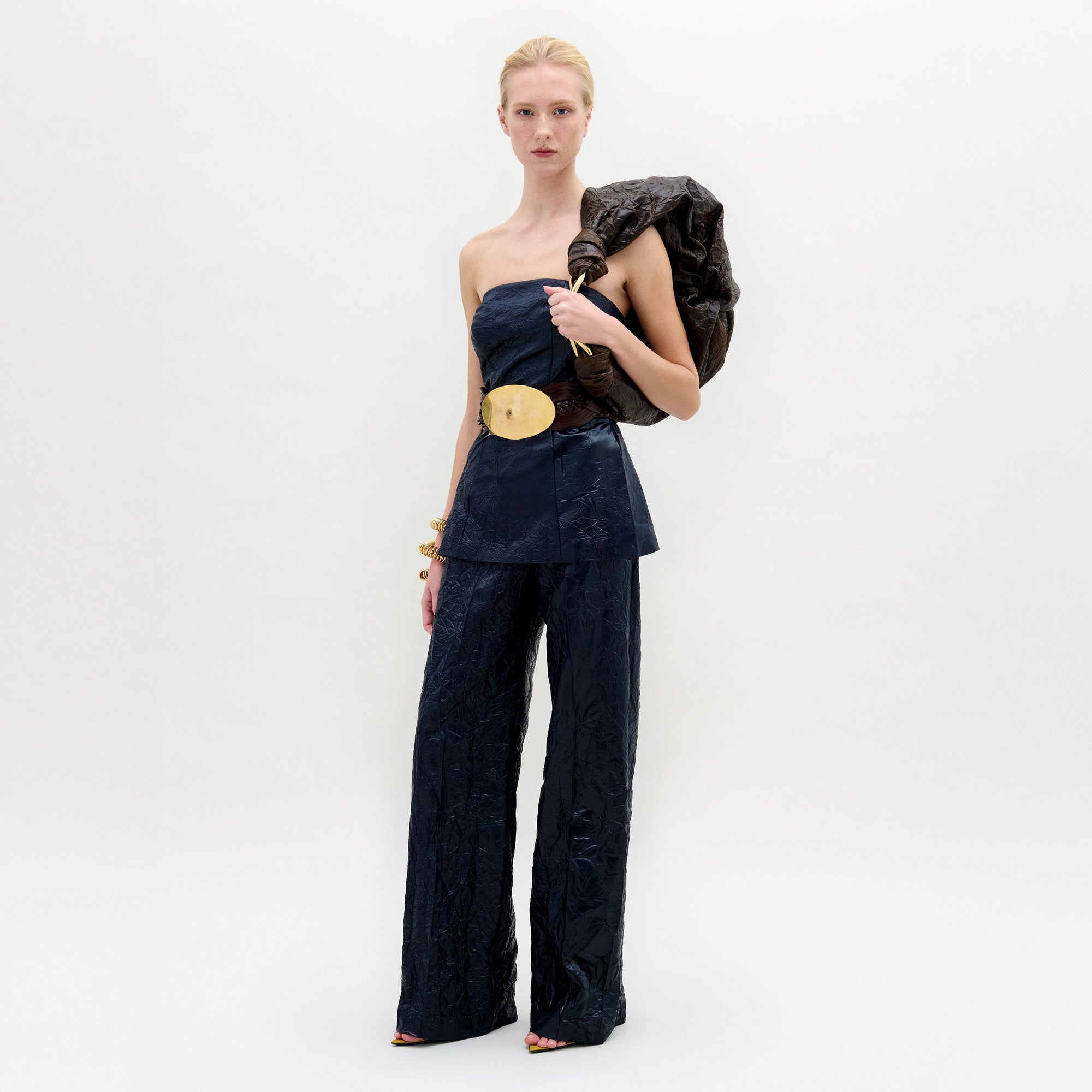 A woman stands against a white background wearing the Emine Pant—a high-waisted, wide-leg silhouette—paired with a dark strapless top, gold belt, bangles, and a textured bag slung over her shoulder.