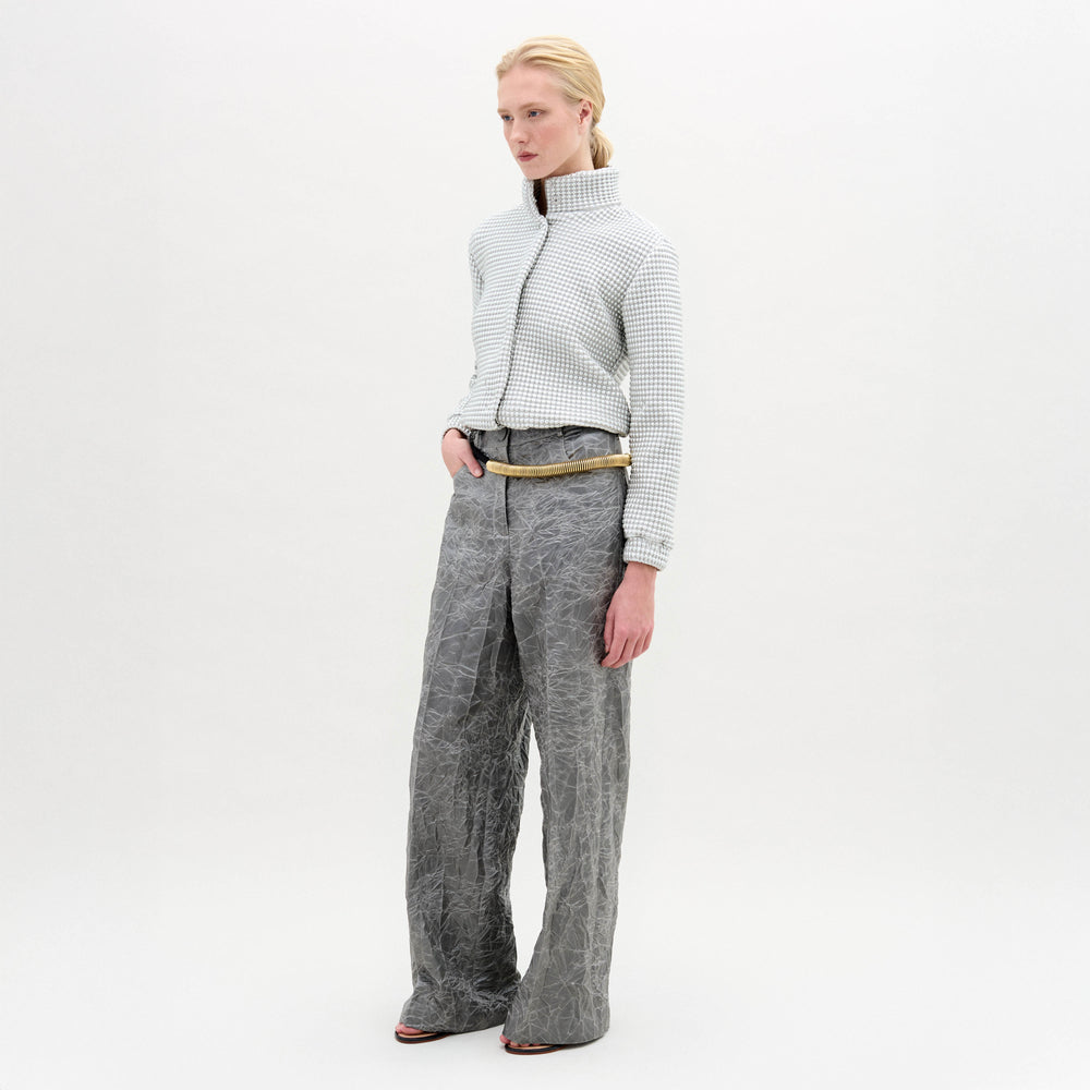 A person stands against a white background, wearing a textured white zip-up jacket and the Emine Pant by SVT DESIGNS—a high-waisted, wide-leg grey pant with a gold belt. Their straight blond hair is pulled back as they look to the side.