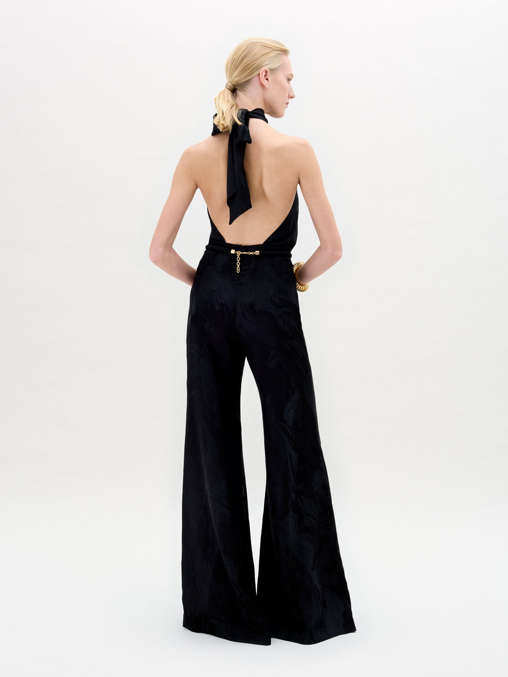 A person stands against a white background in the Enara Jumpsuit, a black halter-neck piece made from luxurious Italian fabric with wide-leg pants, accessorized with elegant gold bangles.