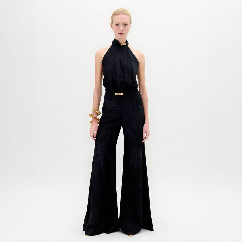 A person stands facing forward against a plain white background, wearing the SVT DESIGNS Enara Jumpsuit—a sleeveless black halter-neck style with wide-leg pants—styled with gold accessories at the wrist and neck.