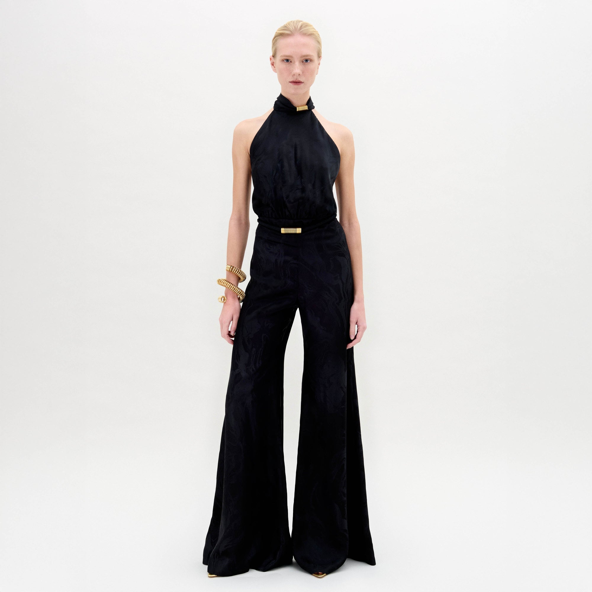 A person stands against a white background in the Enara Jumpsuit, a black halter-neck piece made from luxurious Italian fabric with wide-leg pants, accessorized with elegant gold bangles.