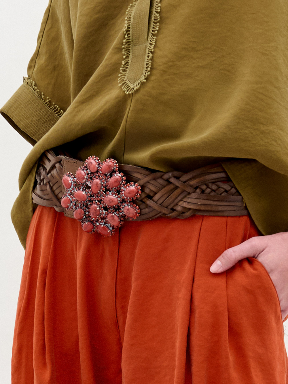 An individual wears an olive green top and orange pants, accessorized with the SVT DESIGNS Eines Belt—a braided leather belt with a large pink bejeweled buckle. One hand rests in their pocket.