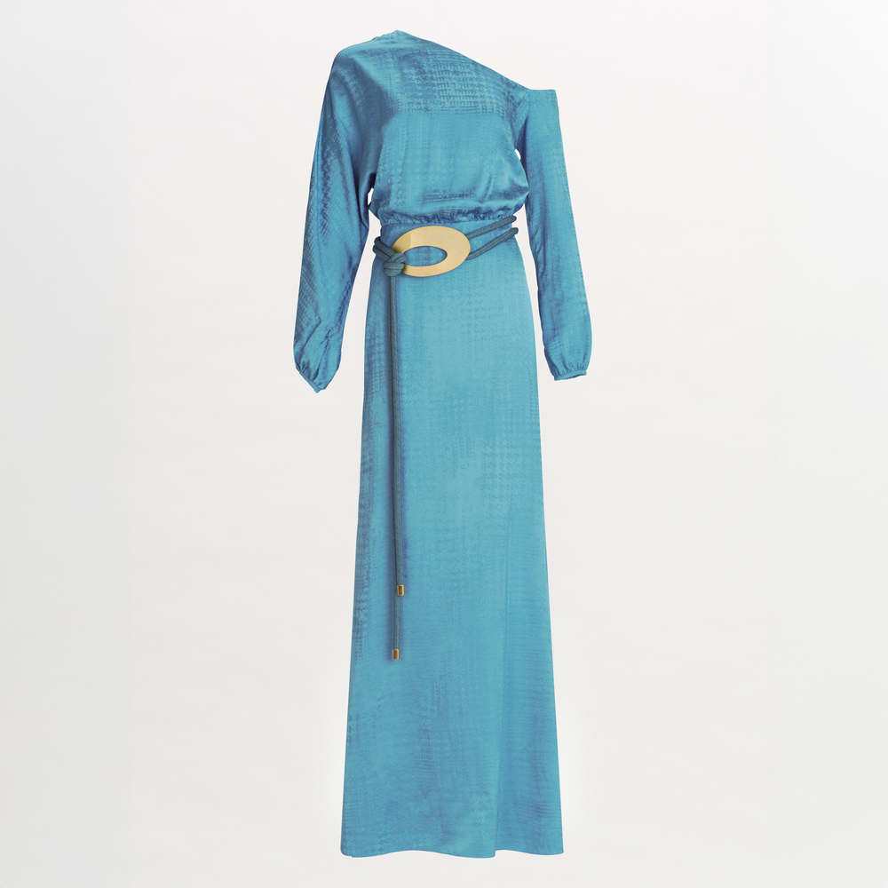 The Elga Dress by SVT DESIGNS is a long, light blue maxi with an asymmetric one-shoulder neckline, long sleeves, subtly textured Italian fabric, and a wide belt featuring a large gold buckle at the waist.