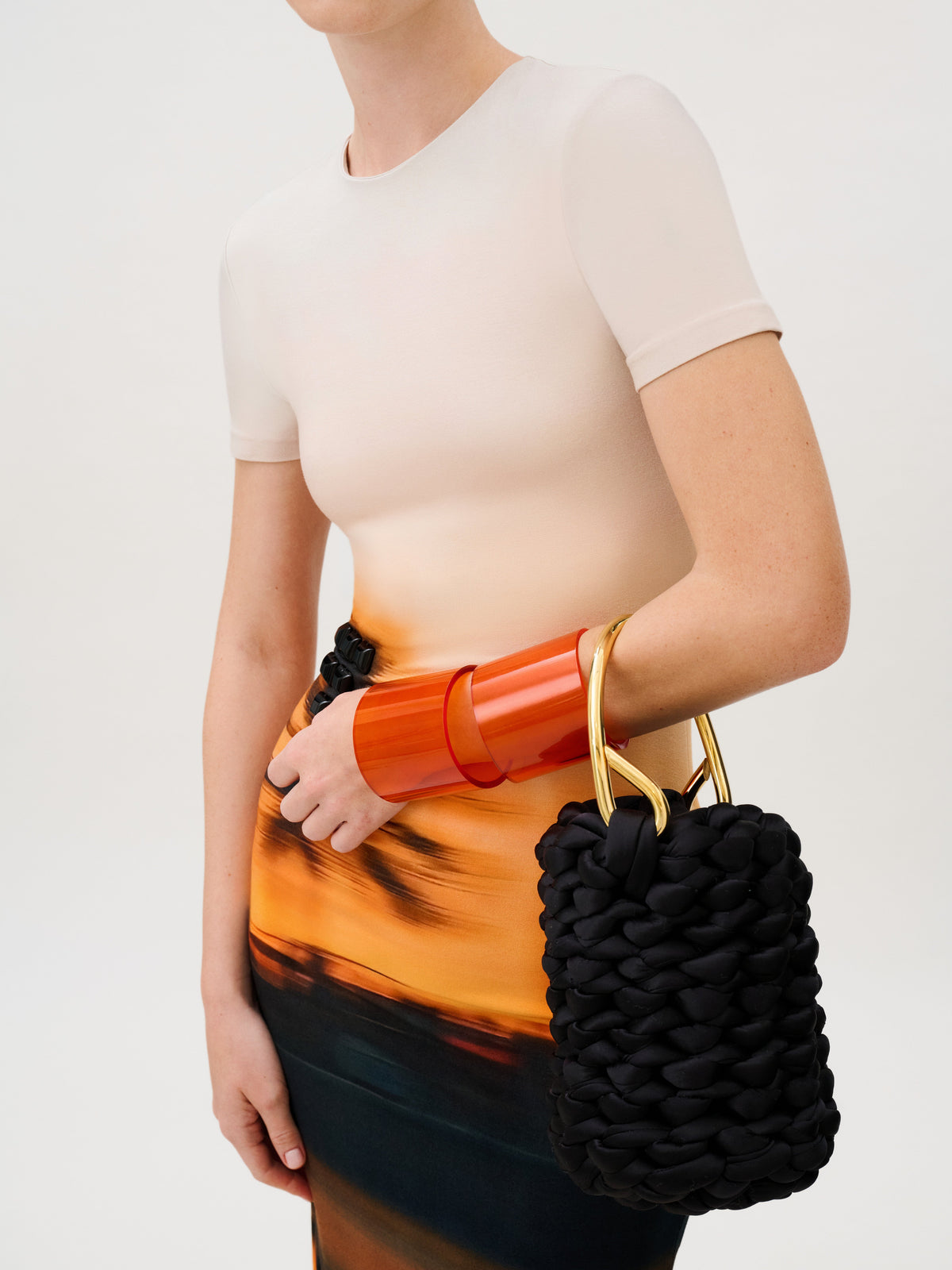 The Elora Dress is a slim fit top in Italian fabric that transitions from beige to an orange and black abstract print, accessorized with a wide translucent orange cuff and a black braided handbag with gold metal handles.