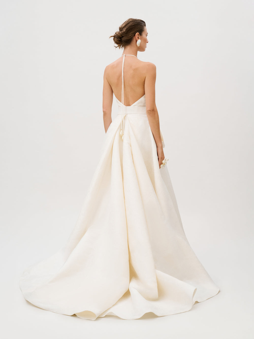 A woman stands facing away, holding a small bouquet, wearing the Elsa Dress—a white halter-neck, backless gown crafted from luxurious Italian fabric with a long train.
