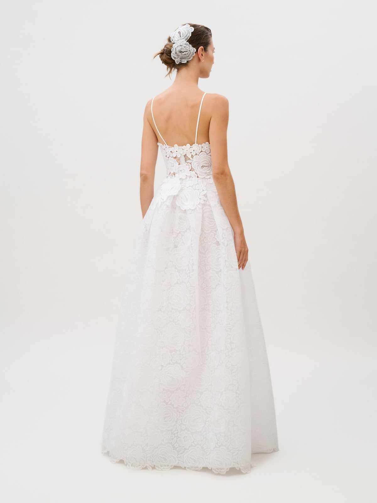A woman stands facing away, highlighting the detailed back of the Esme Dress—a white lace design with thin straps and floral appliqué, meticulously crafted from luxurious Italian fabric. She wears a floral hair accessory. The background is plain and light. Made in Colombia.