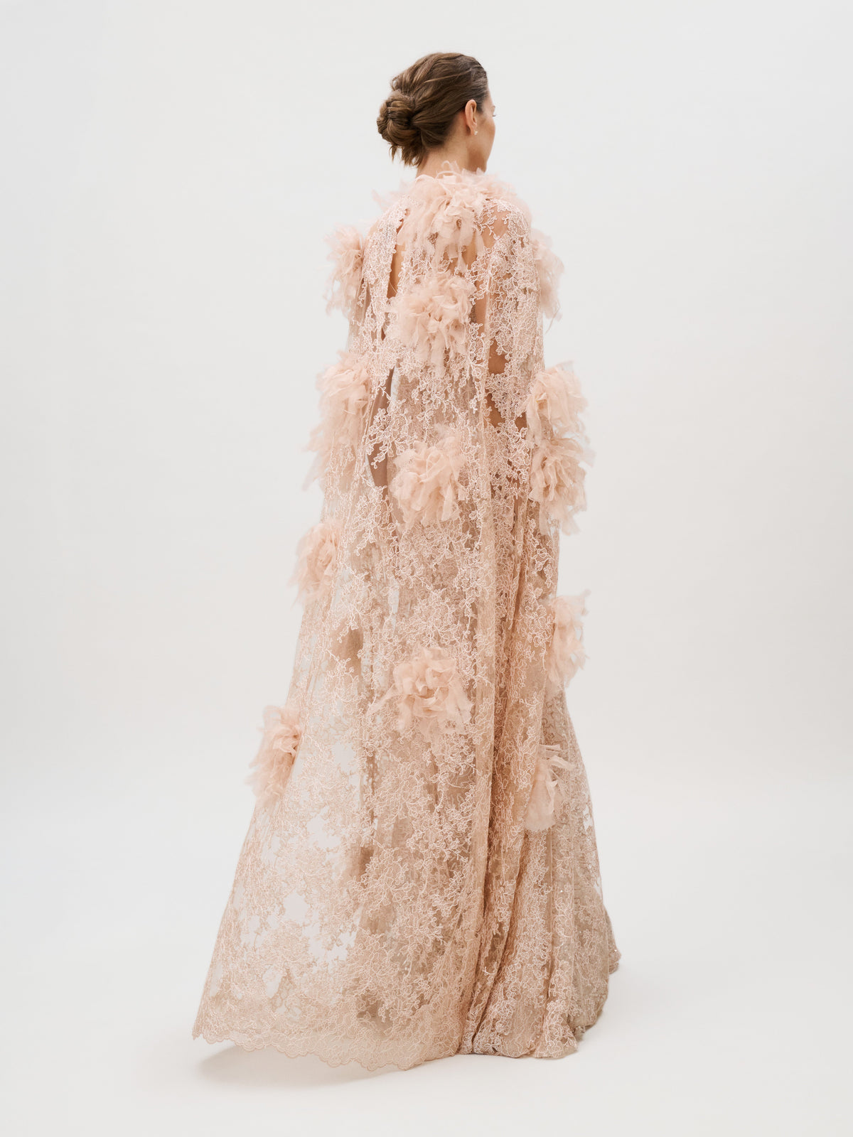 A woman in the Eudora Dress—a floor-length, pale pink lace gown adorned with floral appliqué and crafted from luxurious Italian fabric—stands against a plain white background, facing away from the camera with her hair styled in a low bun.