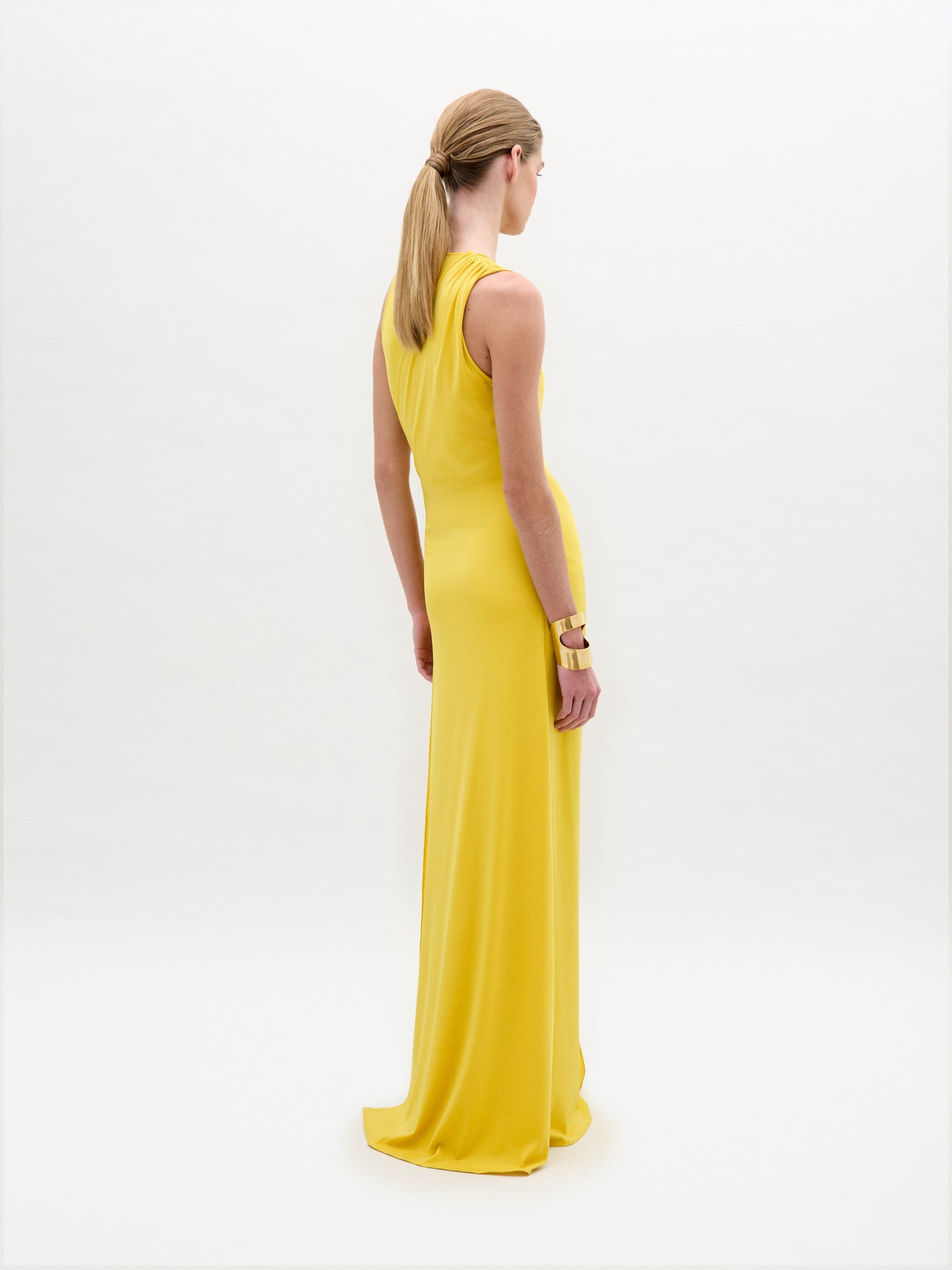 A woman with blonde hair tied back stands facing away, wearing the vibrant yellow Fadila Dress by SVT DESIGNS. The sleeveless maxi dress beautifully fits her silhouette. Her right arm is adorned with gold bangles, set against a plain white background.