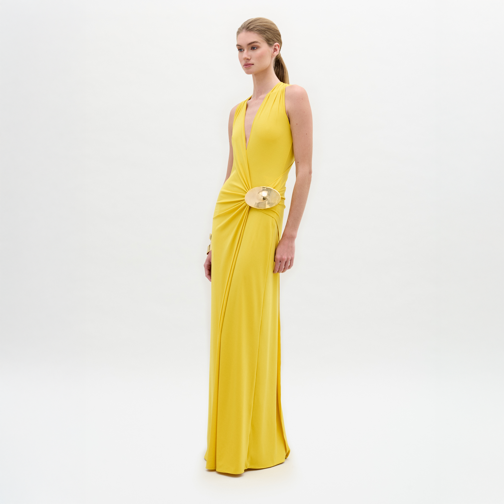 A woman poses against a white background wearing the Fadila Dress by SVT DESIGNS—a sleeveless yellow maxi dress with a deep V-neck, crafted from fine Italian fabric and highlighted by a gold waist embellishment.
