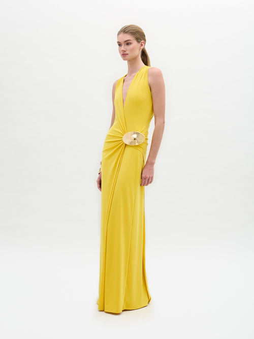 A woman dons the SVT DESIGNS Fadila Dress, a sleeveless yellow maxi with a deep V-neck and fitted silhouette, highlighted by a gold belt. She poses gracefully against a plain white background.