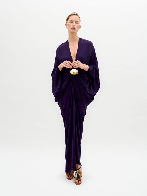 Against a plain backdrop, a person wears the SVT DESIGNS Fiore Tunic, a floor-length, draped dark purple maxi dress with a deep V neckline and an oversized silhouette cinched at the waist with a decorative clasp. Brown sandals with gold-tone embellishments complete the look.